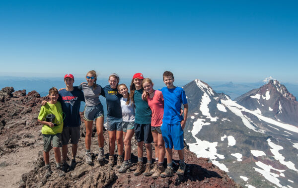 group of kids post hike standing on top of mountain with mountain peaks in background