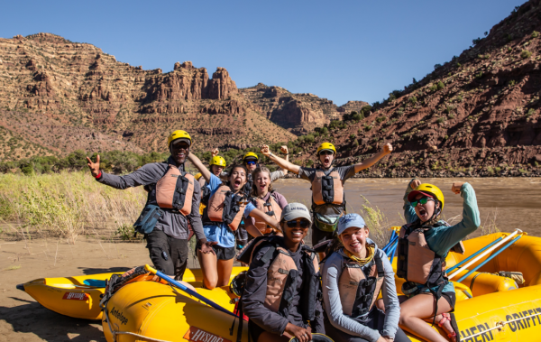 Group of people with their arms in the air on a whitewater raft in the desert.