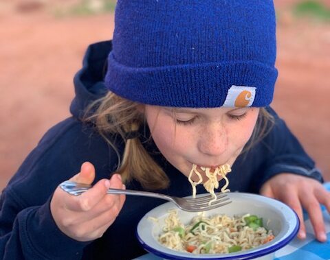 Girl in a blue beanie eating a bowl of ramen noodles in the desert at a picnic table.