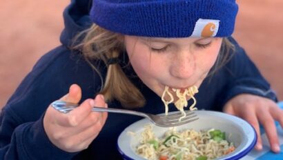 Girl in a blue beanie eating a bowl of ramen noodles in the desert at a picnic table.