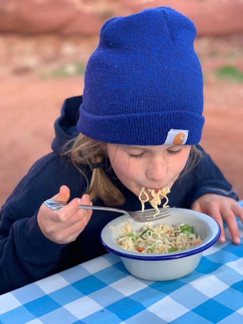 Girl in a blue beanie eating a bowl of ramen noodles in the desert at a picnic table.