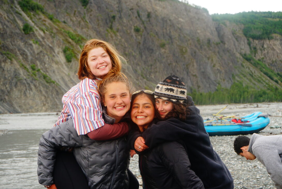 Four girls smiling and hugging next to an alaskan river.