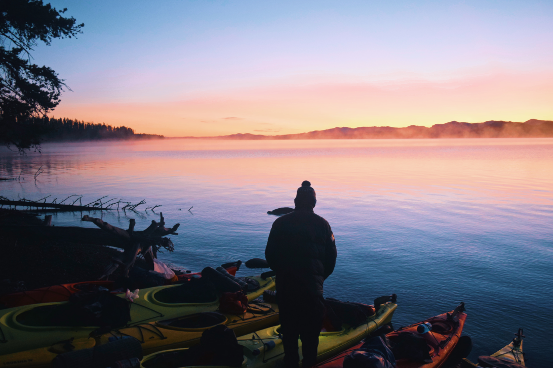 person standing by kayaks on the shore of a lake at sunrise.