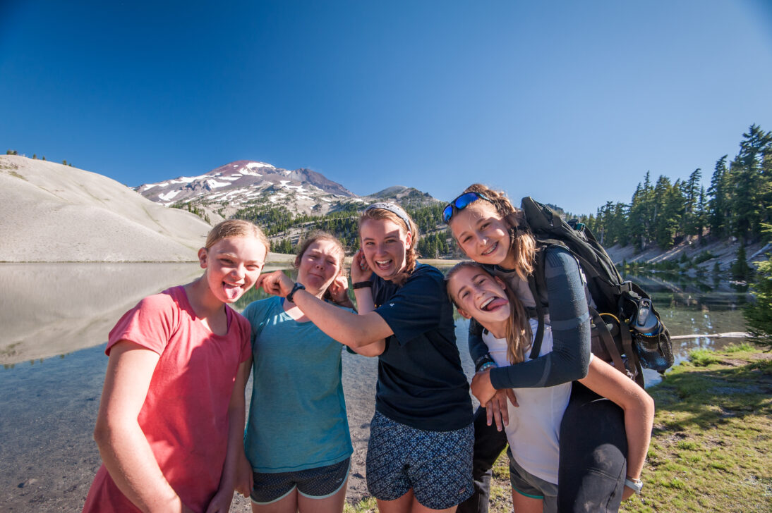 5 girls laughing and being silly for a picture in front of an alpine lake
