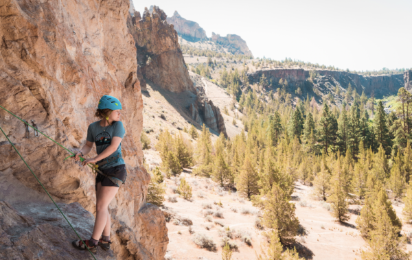 Girl at the top of a rock climbing route looking over her shoulder at vast landscape of pine trees in a valley below her.