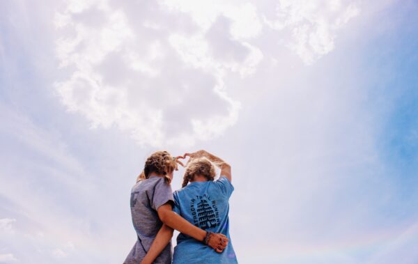 Two girls making a heart with their hands together against a cloudy sky.