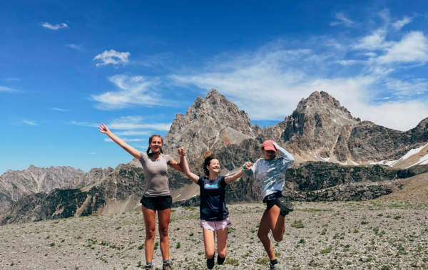 Three girls jumping in the air holding hands in front of a vast mountain on the horizon.