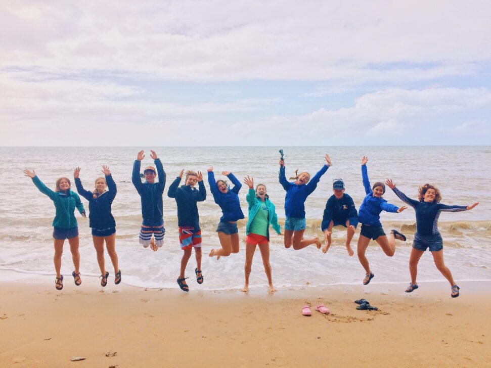 group of students jumping with their hands in the air on the beach.