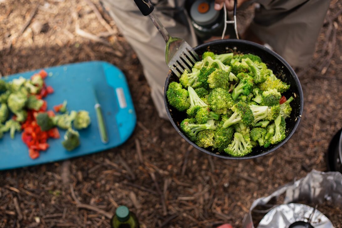 Pan of broccoli being cooked at a campsite with other chopped vegetables.