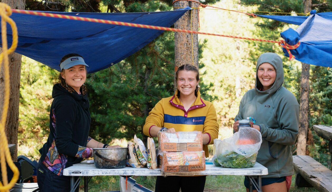 Three people preparing to cook ramen and vegetables under a tarp.