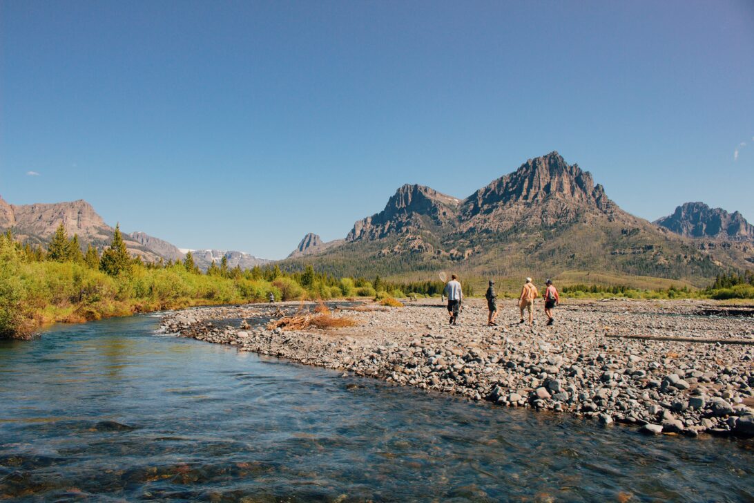 Four people walking on the rocky shore of a river on a sunny day with mountains in the background.