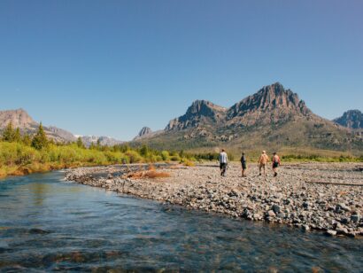 Four people walking on the rocky shore of a river on a sunny day with mountains in the background.