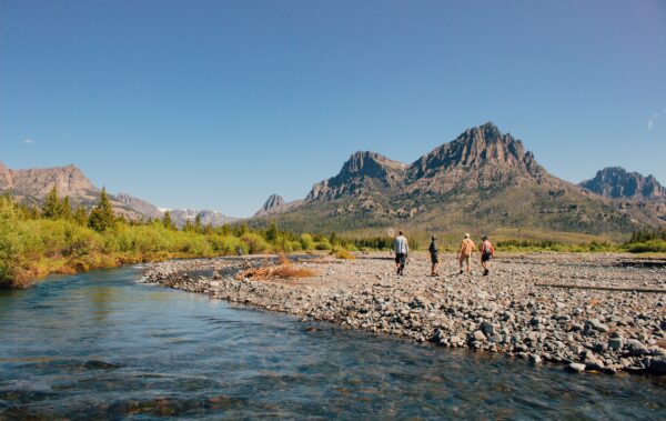 Four people walking on the rocky shore of a river on a sunny day with mountains in the background.