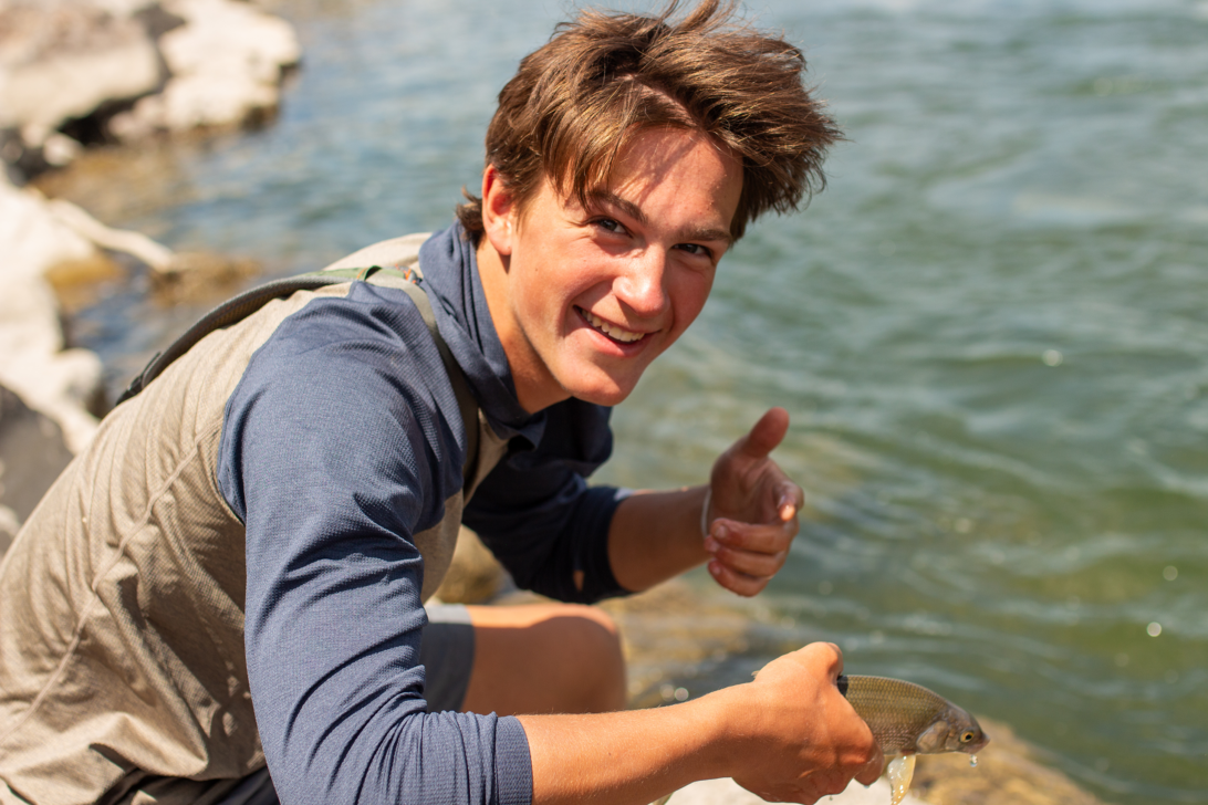 Student holding a fish in one hand and a thumbs up with his other hand by a river.
